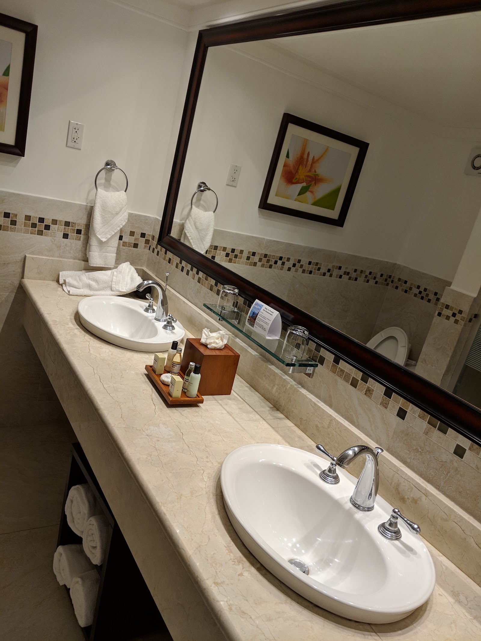 Bathroom vanity with two white oval sinks, mosaic tile accent, framed flower artwork, and white towels hung on chrome rings.