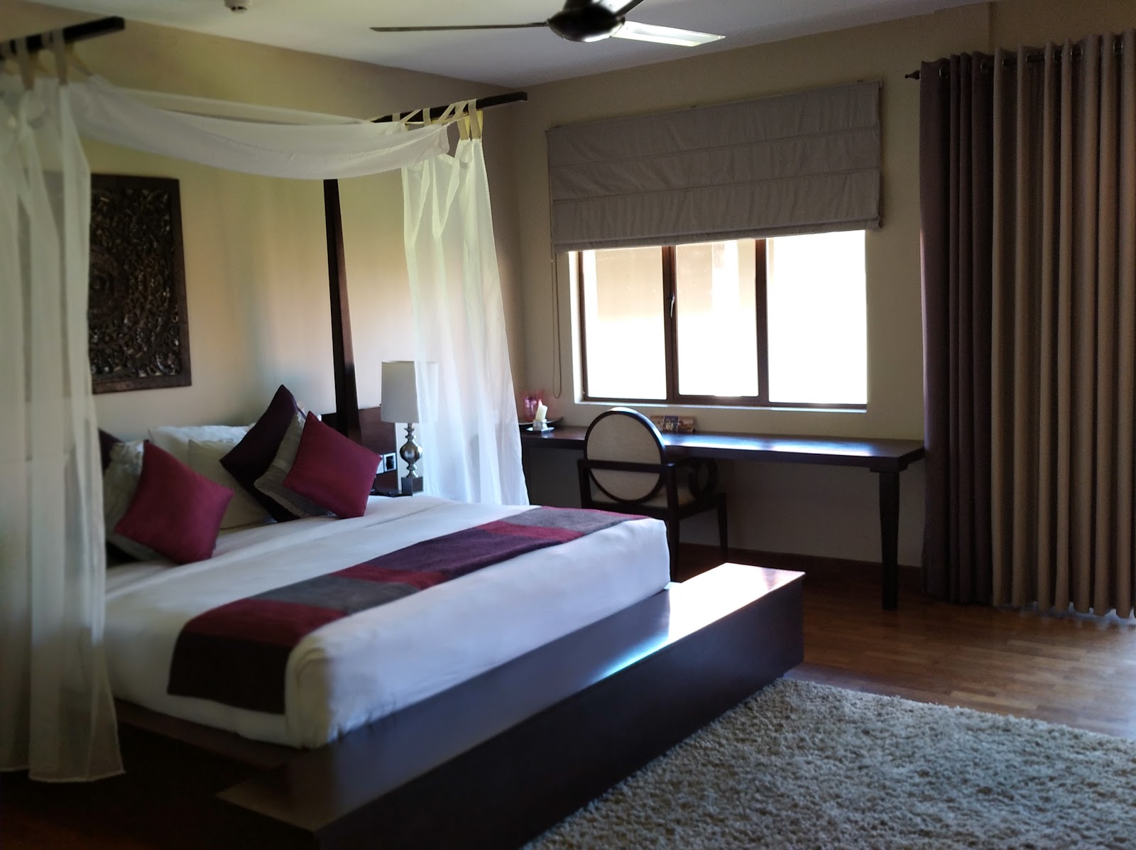 Resort guestroom with four-poster bed draped in sheer white curtains, burgundy accent pillows, work desk by windows, and dark wood furnishings.