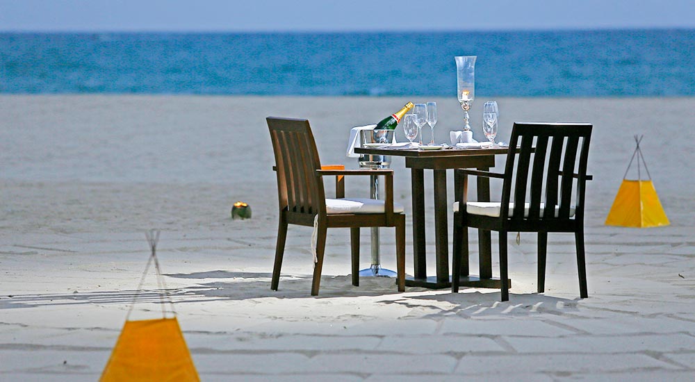 Two black chairs face a table set with champagne and glasses on a sandy beach overlooking turquoise ocean water.