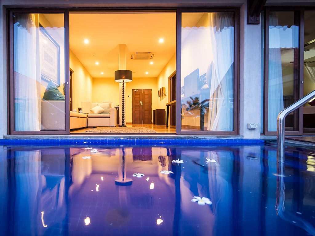 Illuminated resort suite with large glass doors overlooking a blue pool at dusk, cream sofa and wooden entrance visible inside.