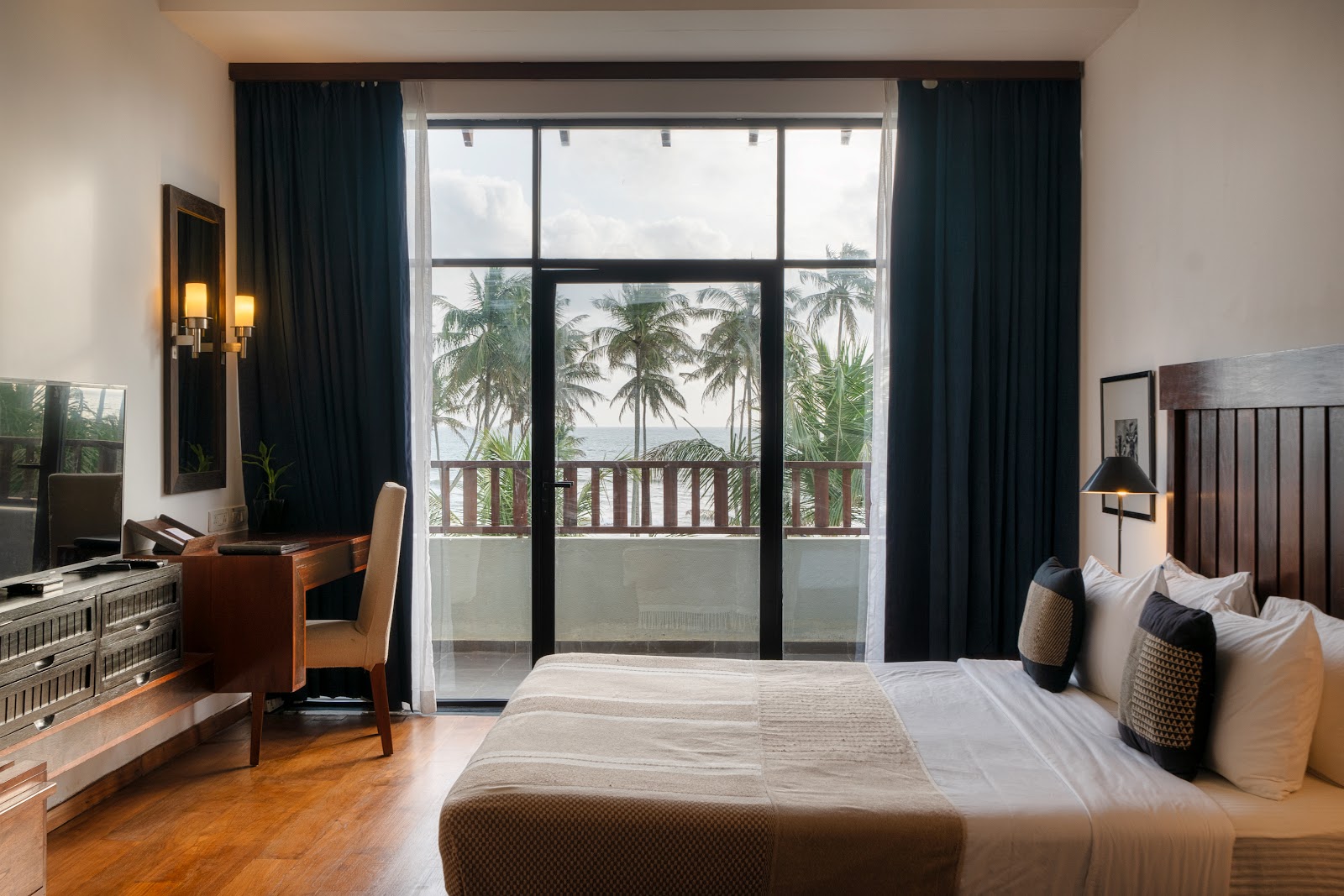 Hotel room with bed, desk, and large windows framed by dark curtains overlooking palm trees and ocean at The Blue Water Hotel and Spa.