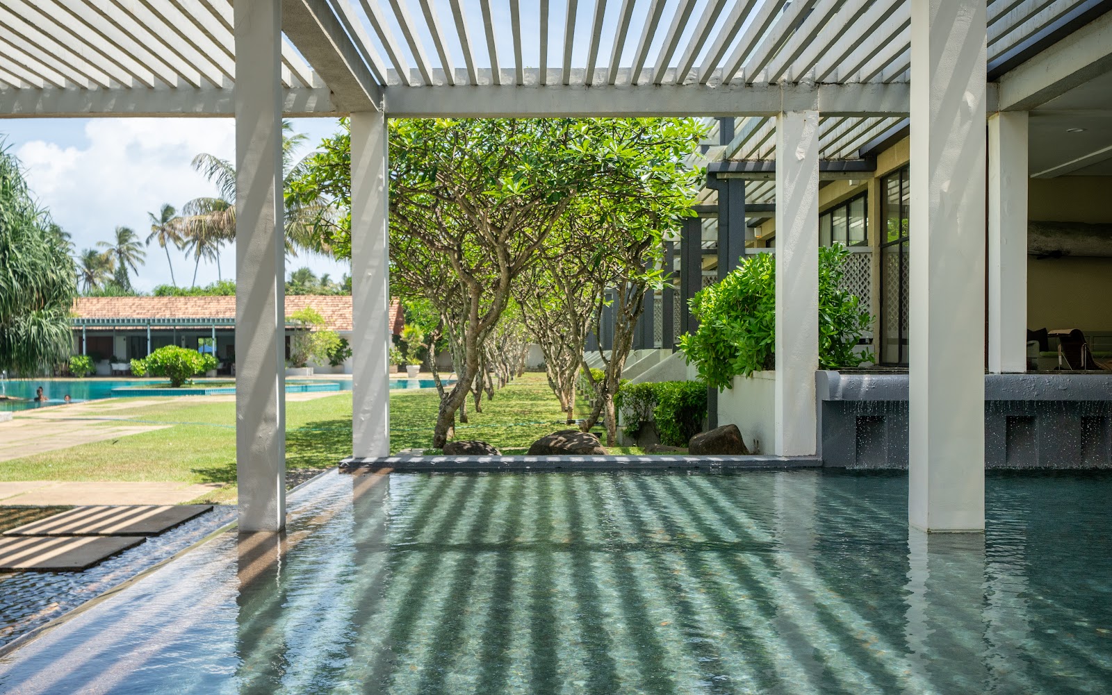 Covered infinity pool at The Blue Water Hotel and Spa with pergola, mature trees, and manicured lawn beyond.