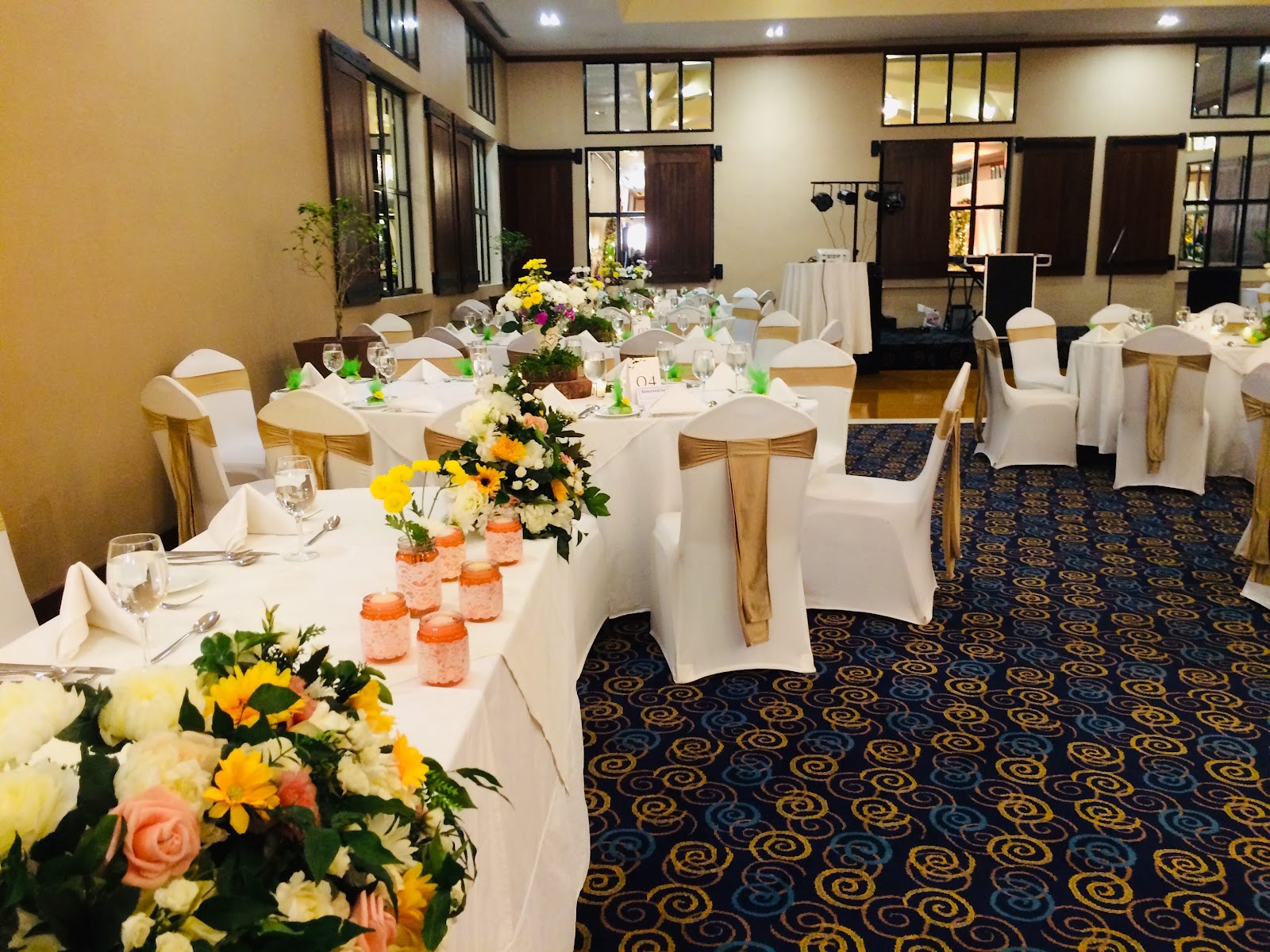 Banquet hall with long tables dressed in white linens, wooden chairs with gold sashes, and colorful flower centerpieces in yellow, pink and white.
