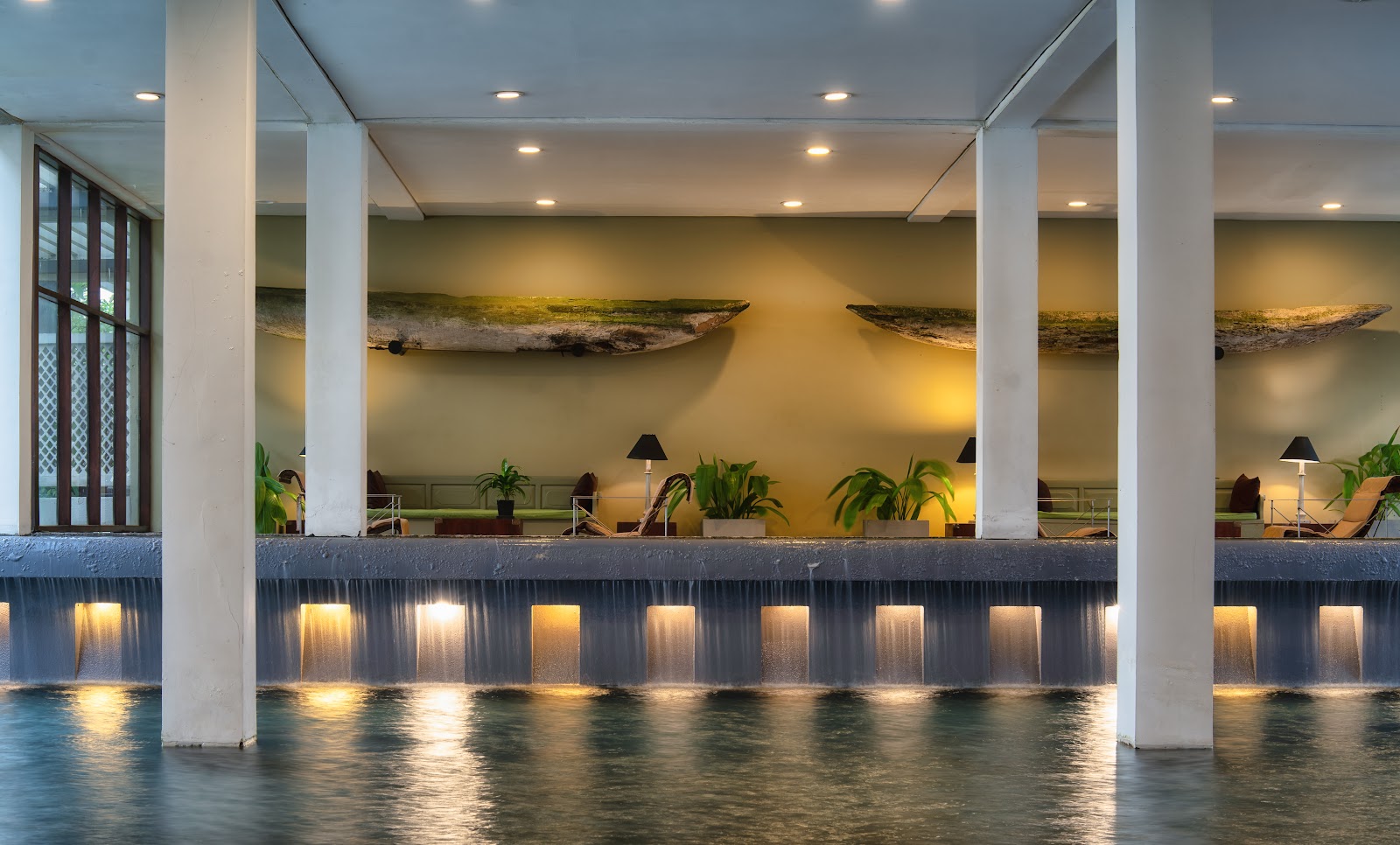 Illuminated waterfront lounge at The Blue Water Hotel and Spa with white columns, recessed lighting, driftwood wall art, potted plants, and shallow water reflecting the warm glow.