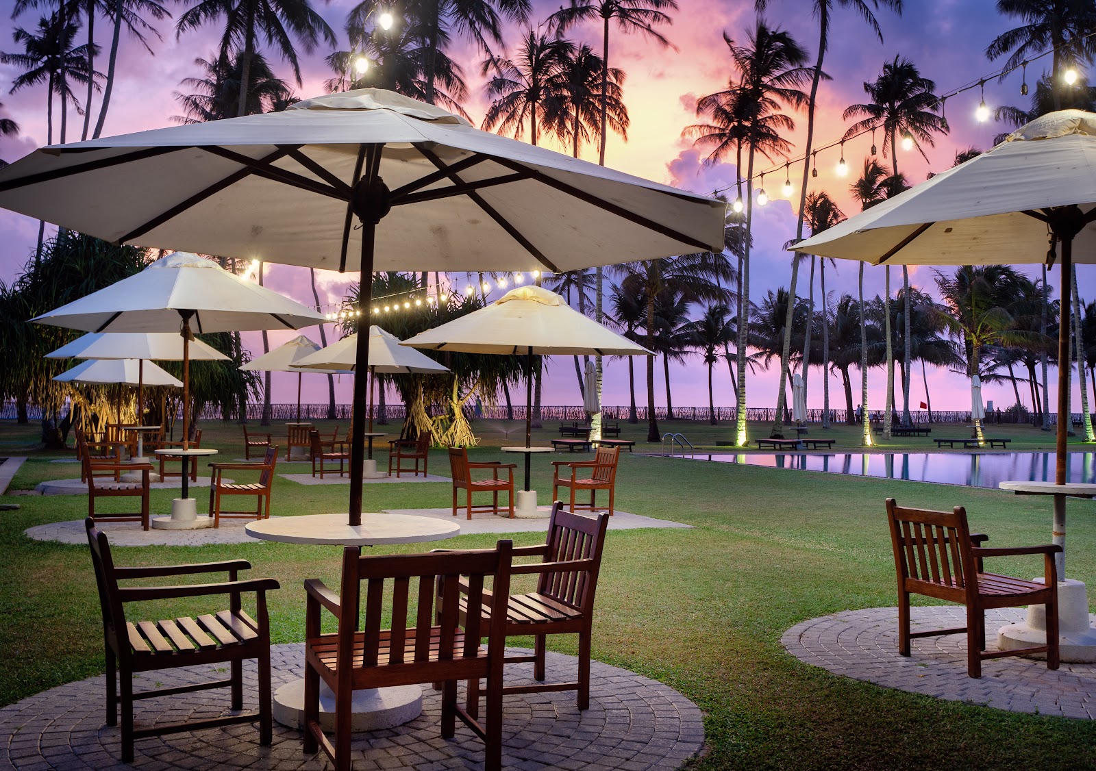 Multi-story colonial-style resort building with terracotta roof beside lagoon, palm trees, white umbrellas, and manicured lawn under blue sky.