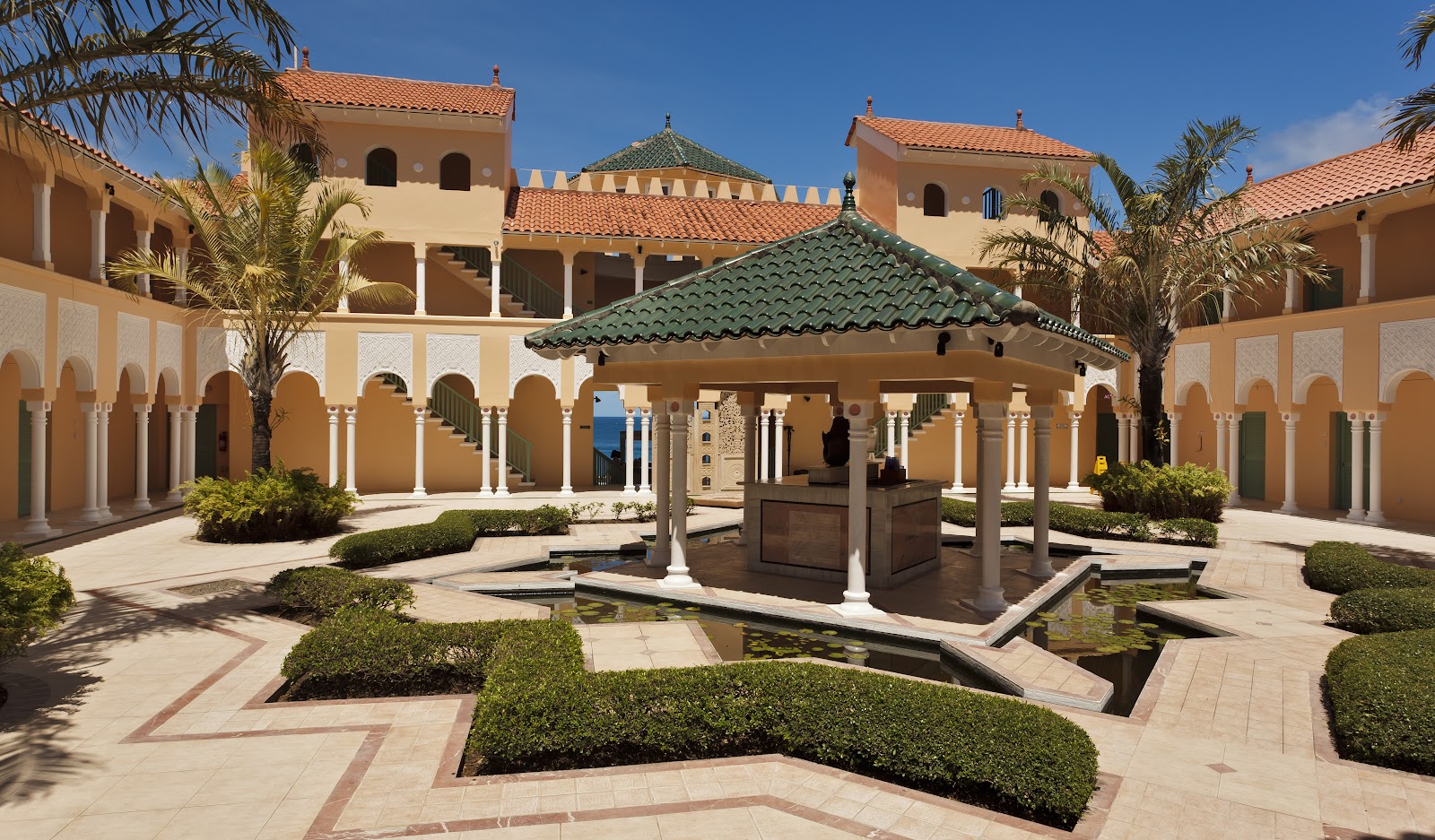 Moroccan-style courtyard with arched colonnades, a central pavilion with green tile roof, manicured hedges, reflecting pool, and palm trees under clear blue sky.