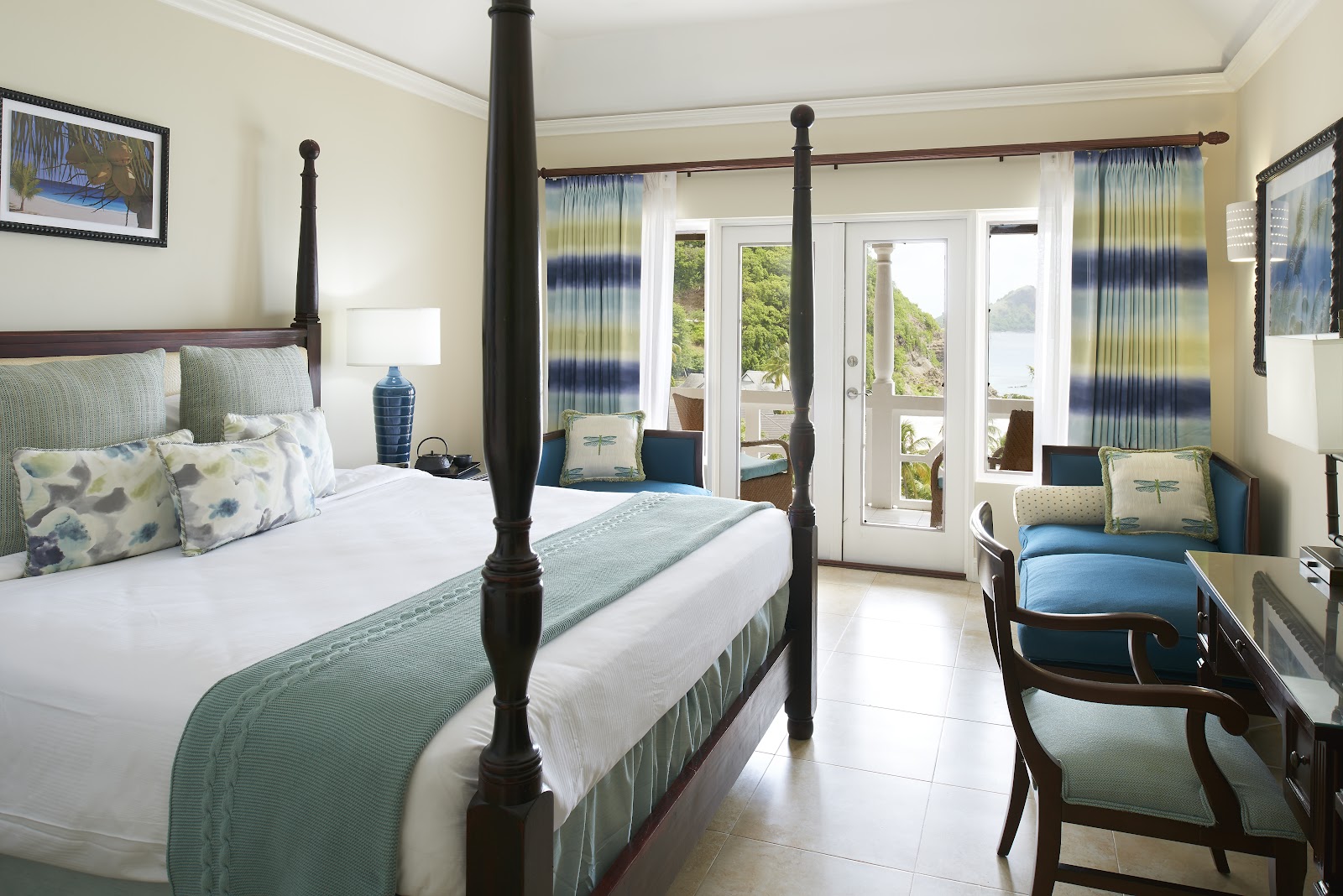 Resort bedroom with four-poster bed, ocean view through glass doors opening to a balcony with green hillside backdrop.