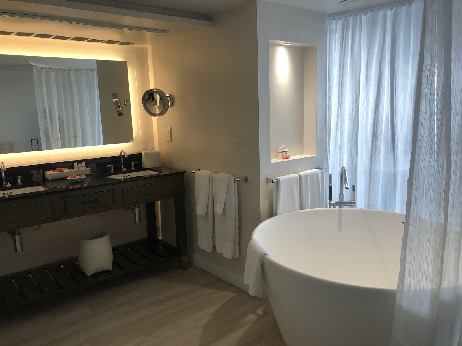 Modern bathroom with freestanding white tub, dark vanity with lighted mirror, and sheer curtains at The BodyHoliday.