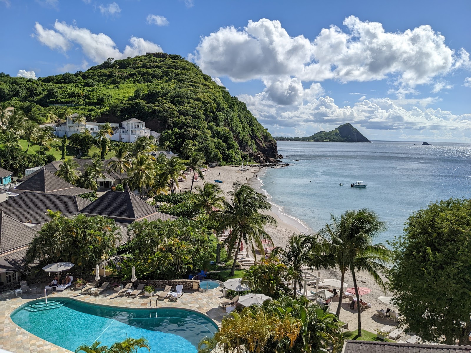 Resort pool and beach with lush green hillside, white colonial buildings, palm trees, and turquoise Caribbean waters at The BodyHoliday.