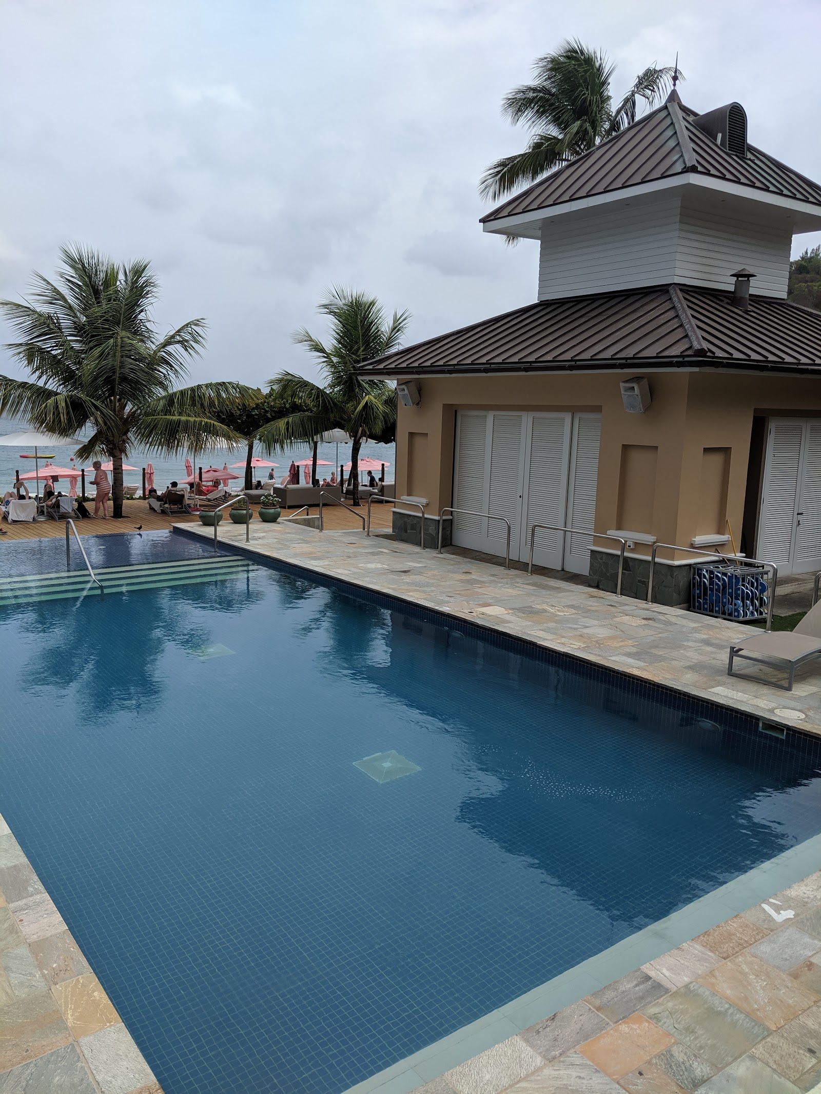 Resort pool with metal-roofed cabana and beach beyond, palm trees framing the scene and ocean in the distance.