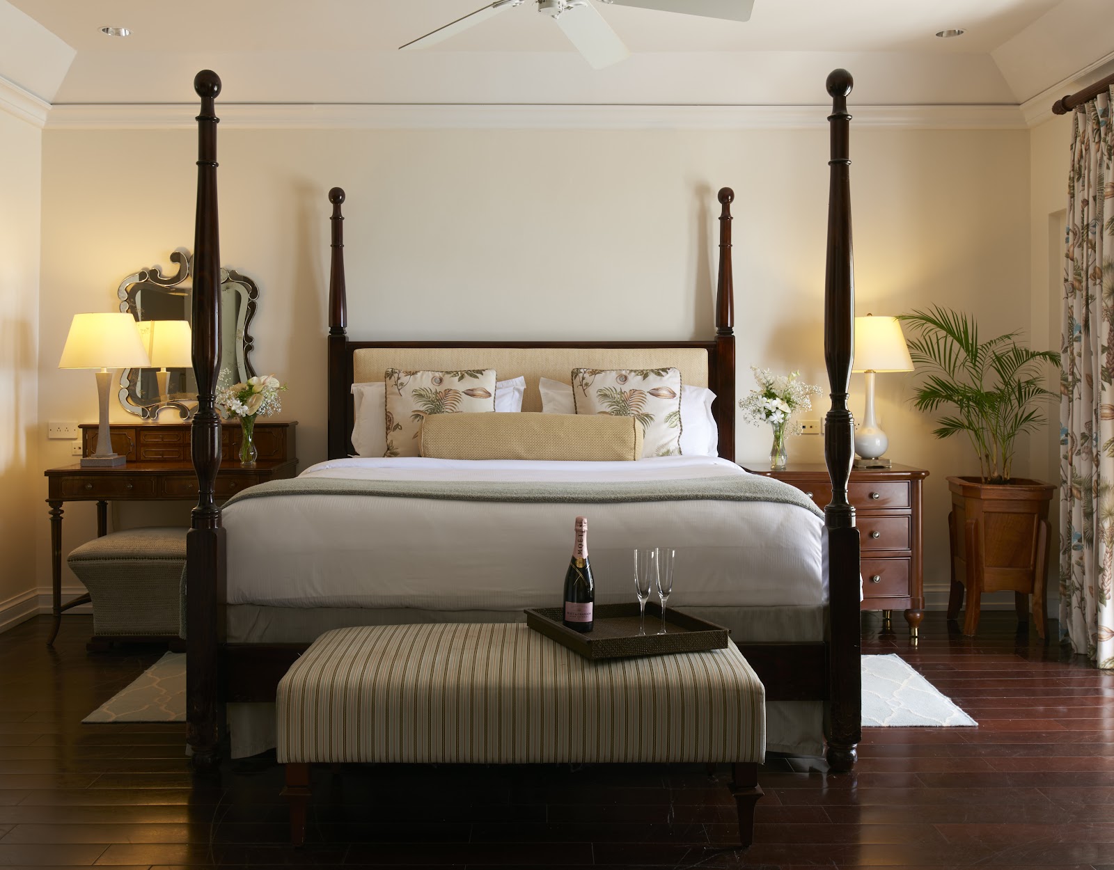 Four-poster bed with dark wood frame in a cream-colored resort bedroom with matching nightstands, lamps, and a striped ottoman at the foot of the bed.