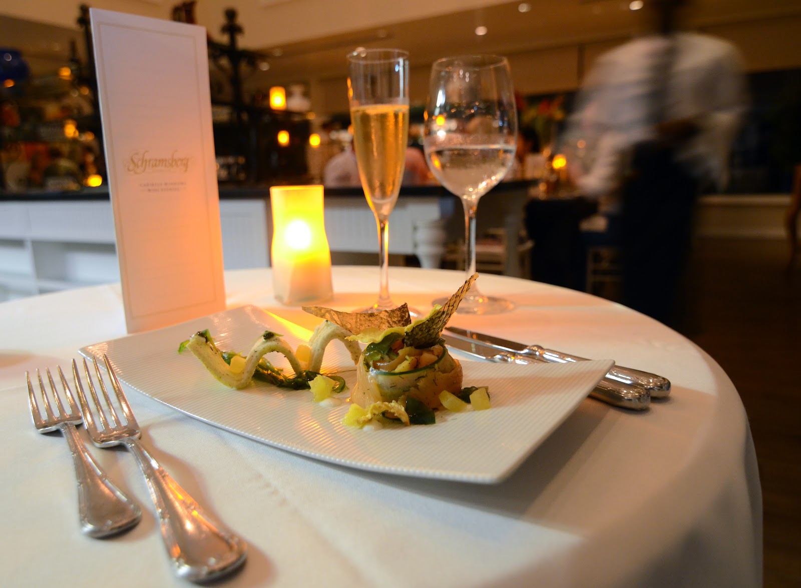 Plated appetizer with vegetables and crispy garnish beside champagne and water glasses on white tablecloth at The BodyHoliday dining room.
