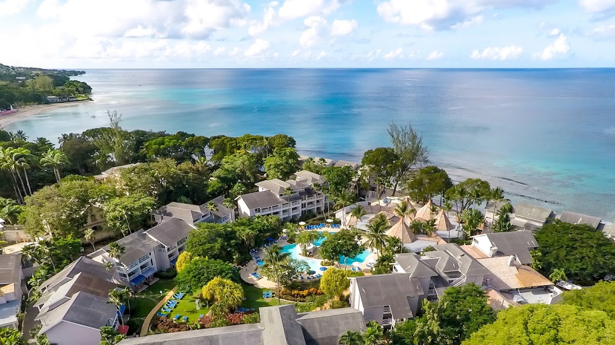 The Club Barbados Resort & Spa sits on a tropical beachfront with turquoise waters, featuring pool areas, palm trees, and white buildings nestled among lush green grounds.