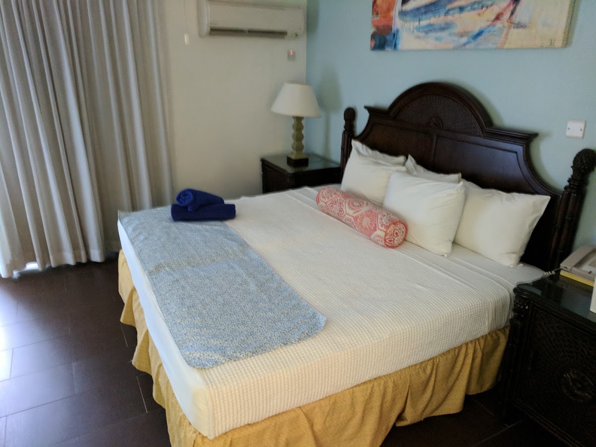 Hotel bedroom with dark wood headboard, white pillows, cream bedspread, and gold bed skirt at The Club Barbados Resort & Spa.