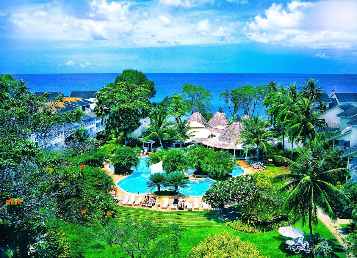 Aerial view of The Club Barbados Resort & Spa with turquoise pool, thatched-roof buildings, lush tropical grounds, and deep blue ocean beyond.