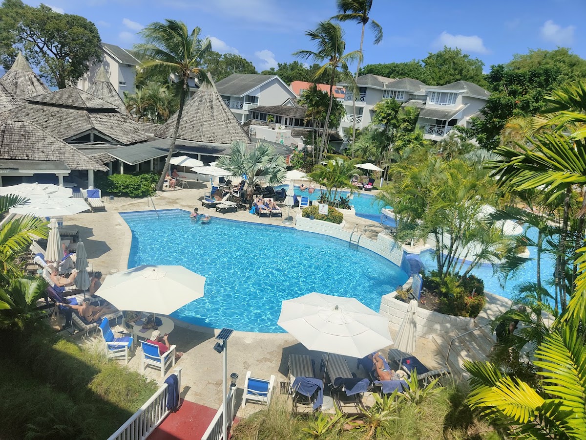 Guests relax by the main pool at The Club Barbados Resort & Spa, surrounded by palm trees and residential buildings under clear skies.