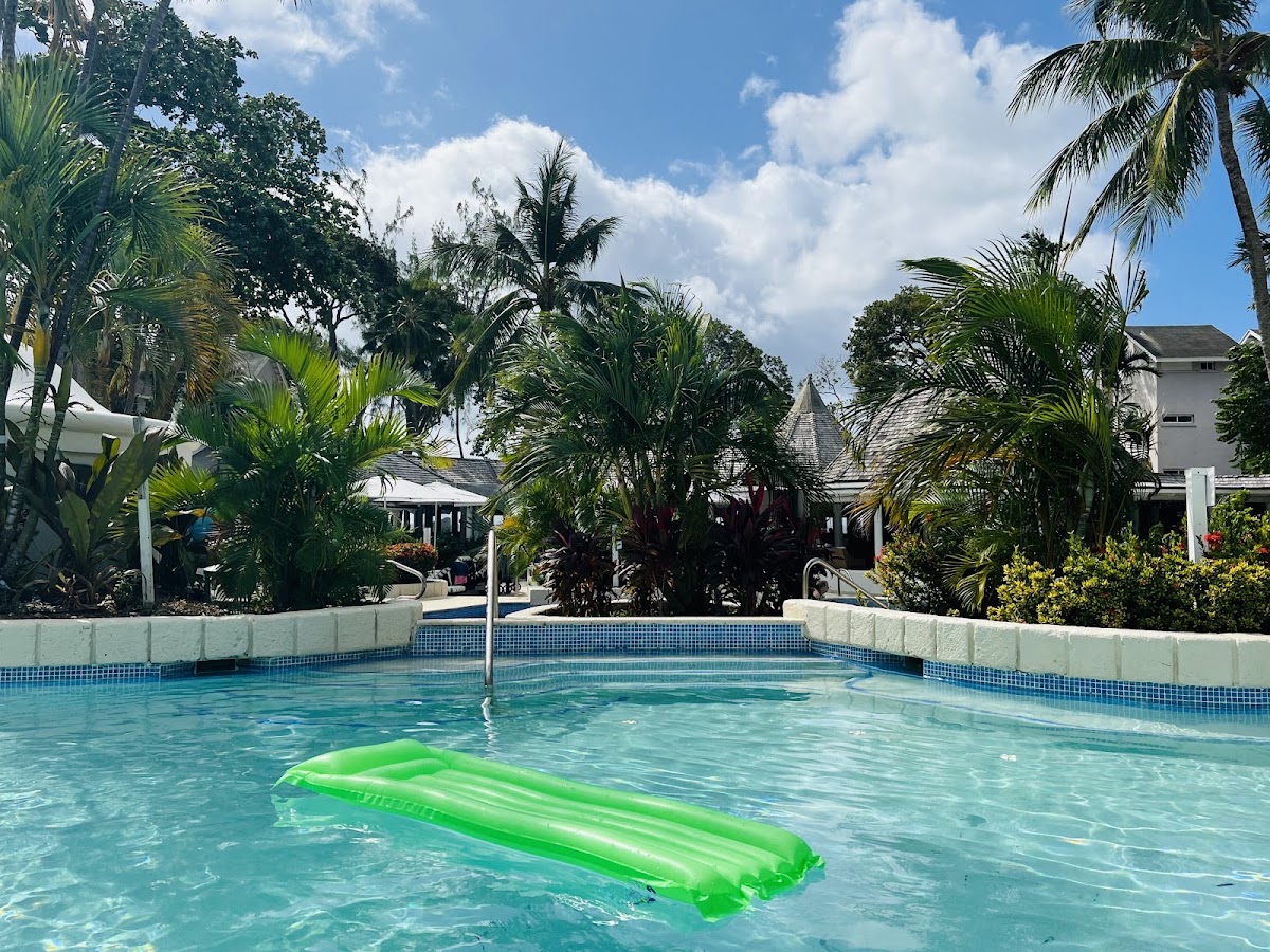 Green inflatable float in a clear turquoise pool at The Club Barbados Resort & Spa, surrounded by palm trees and resort buildings.
