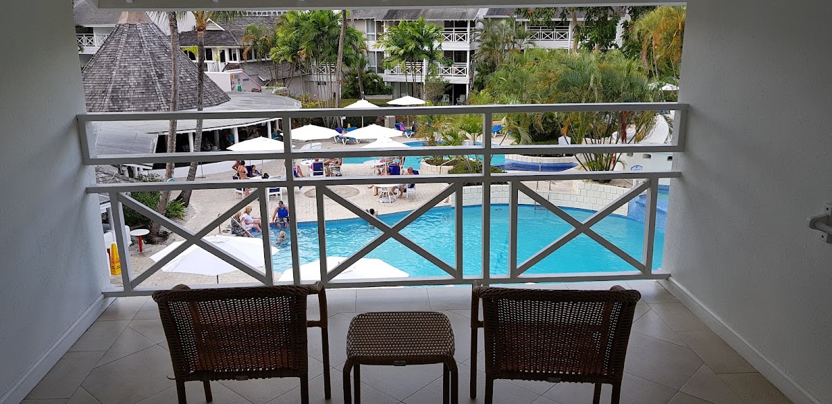 View from a room balcony at The Club Barbados Resort & Spa with two wicker chairs overlooking the bright blue swimming pool and landscaped grounds below.