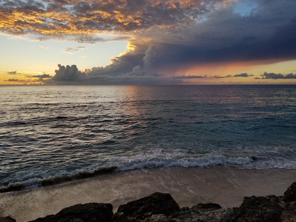 Sunset over the ocean at The Club Barbados Resort & Spa, with dramatic storm clouds lit in gold and orange, waves rolling onto sandy beach.
