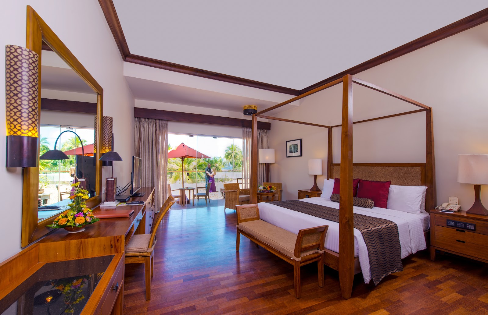 Spacious resort suite with wooden four-poster bed, vanity desk, and balcony overlooking tropical gardens and red umbrellas.
