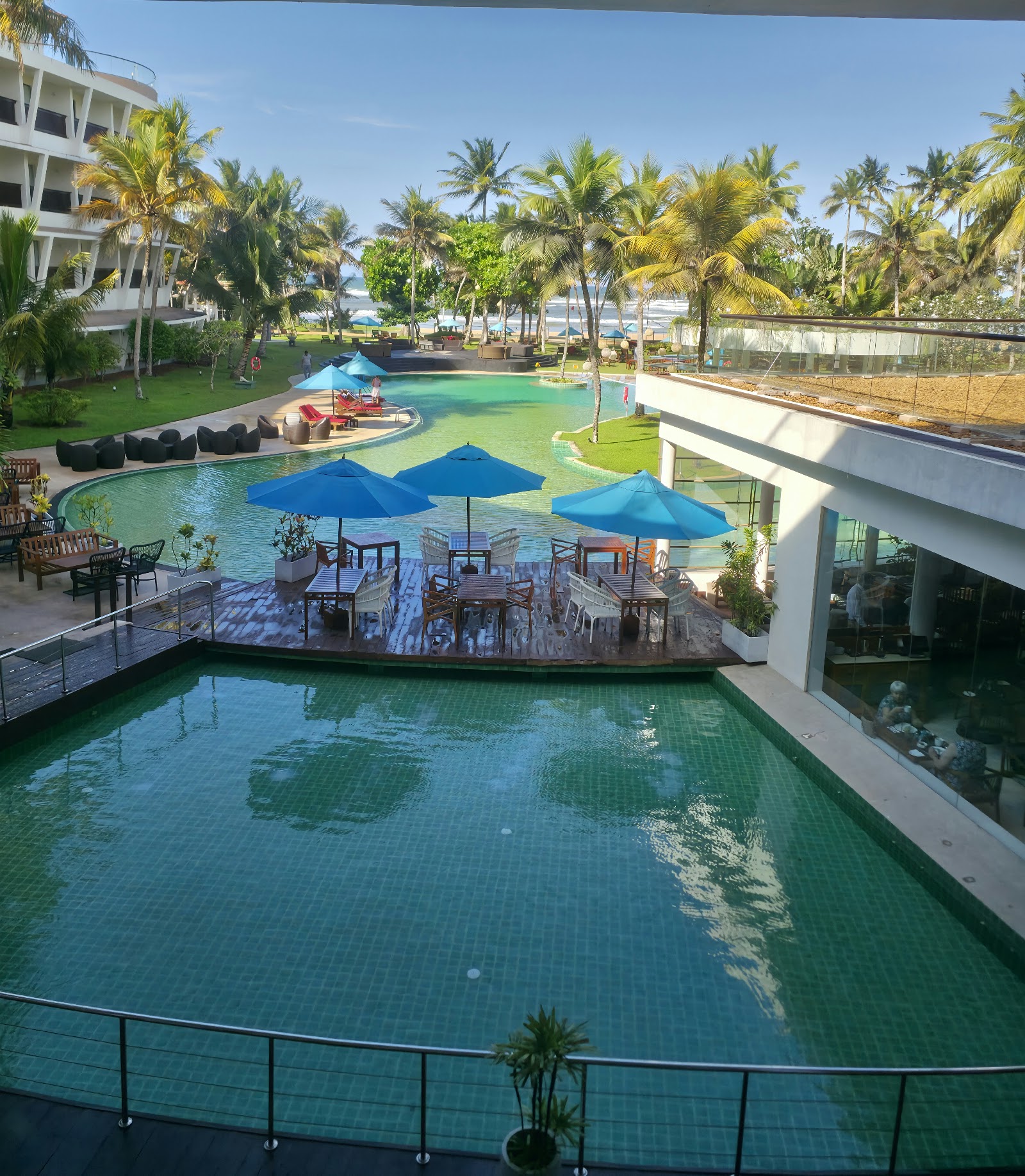 Resort pools and grounds at The Eden Beruwala, with blue umbrellas, palm trees, and ocean view beyond.