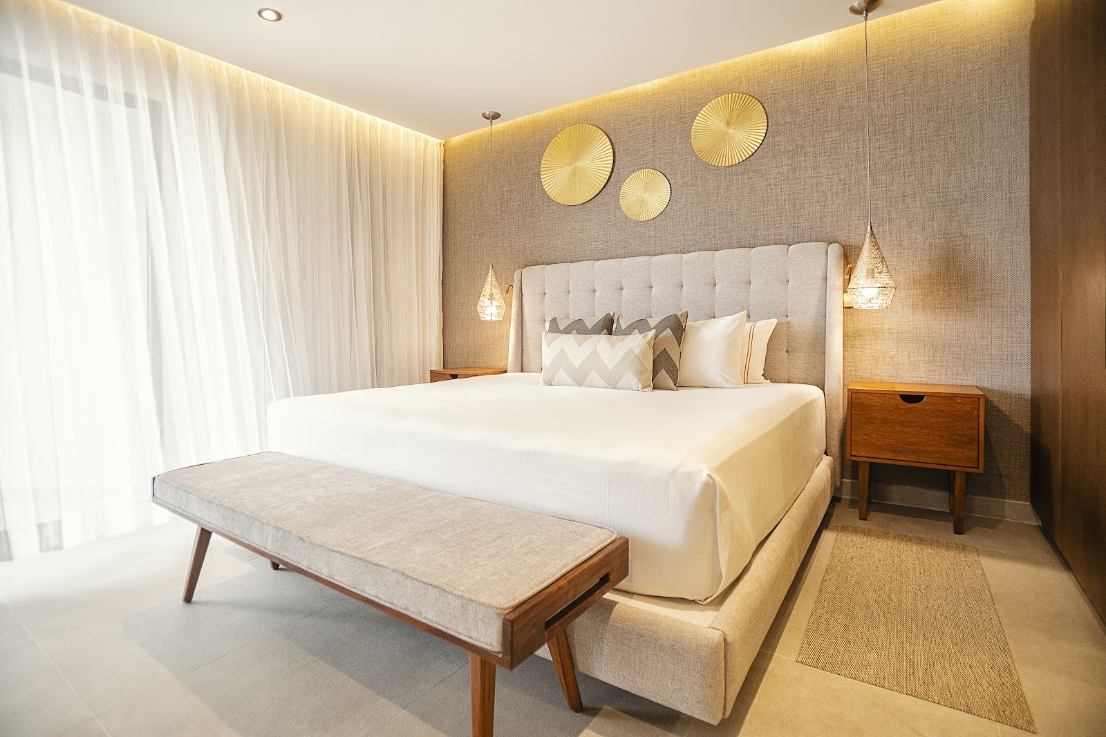 Modern resort bedroom with cream upholstered bed, gold pendant lights, textured walls, and neutral furnishings at The Fives Oceanfront Riviera Maya.