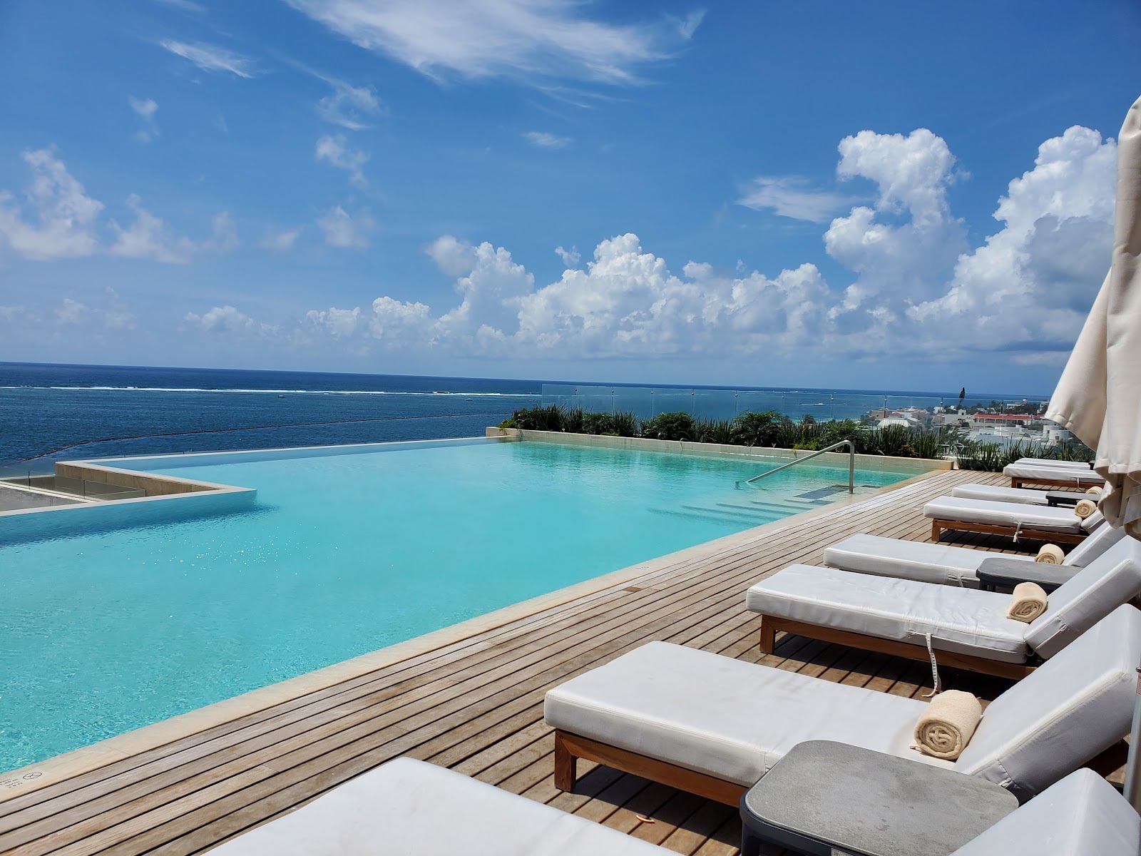 Infinity pool with turquoise water overlooking the Caribbean Sea, wooden deck lined with white lounge chairs and umbrellas under blue sky.