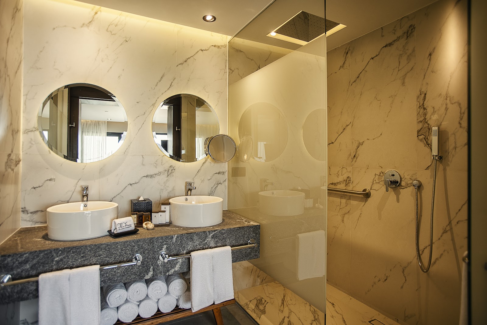 Luxury bathroom with marble walls, dual round vessel sinks, illuminated mirrors, and walk-in shower with glass enclosure at The Fives Oceanfront.