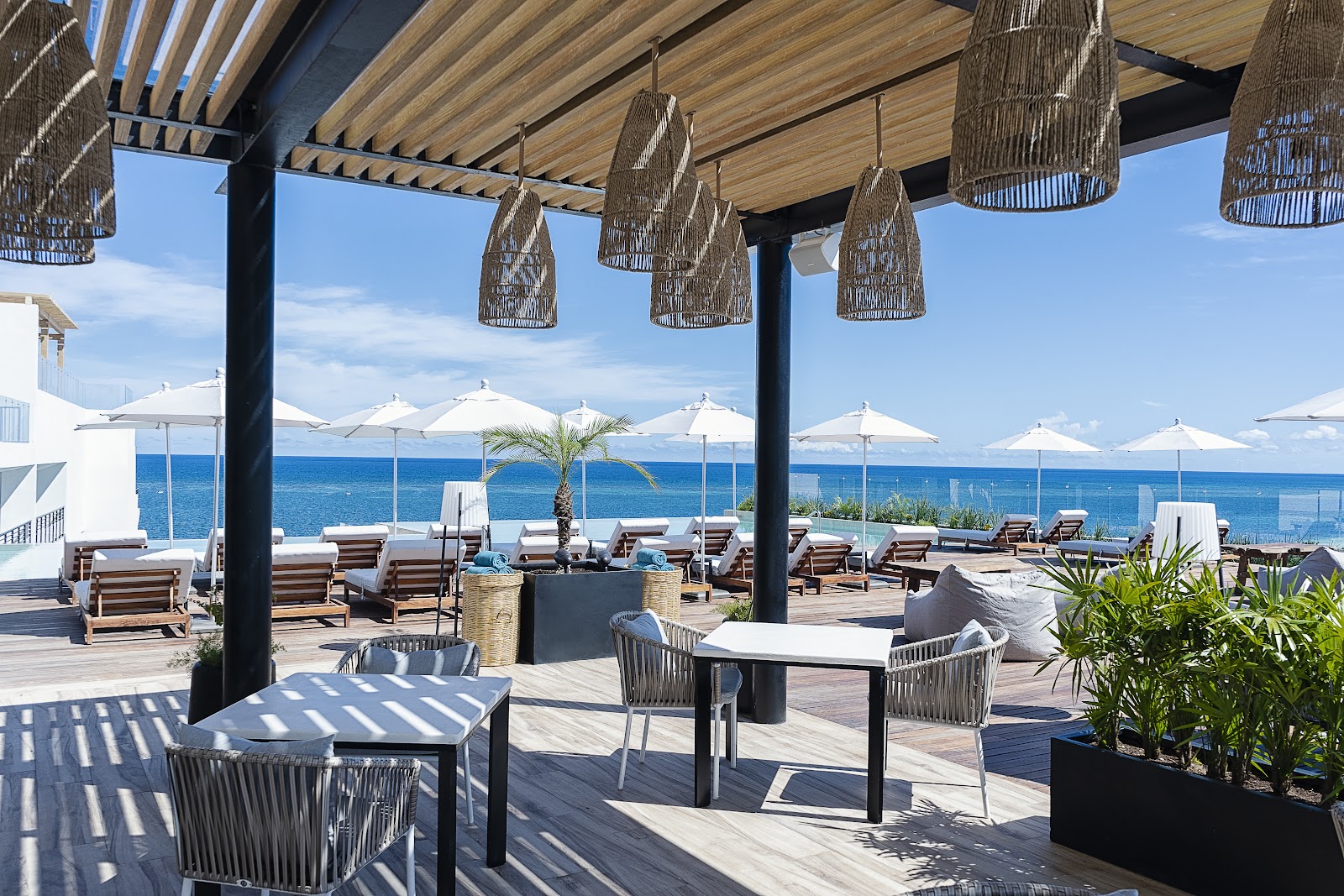 Oceanfront terrace with woven pendant lights over dining tables, lounge chairs, white umbrellas, and views of blue sea and sky.