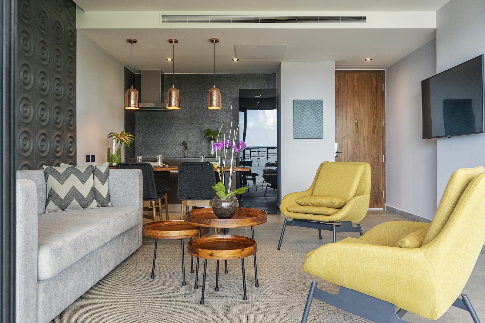 Modern suite living area with gray sofa, yellow chairs, wooden side tables, copper pendant lights, and kitchenette with dark backsplash.