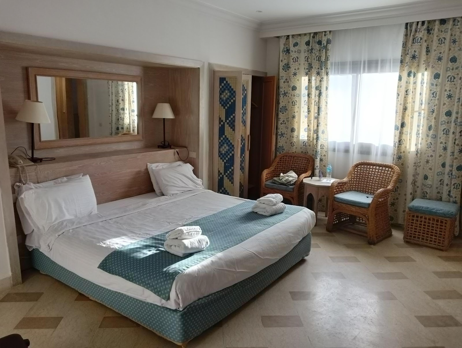 Hotel bedroom with queen bed, wooden headboard with mirror, wicker chairs, and patterned curtains overlooking a window.