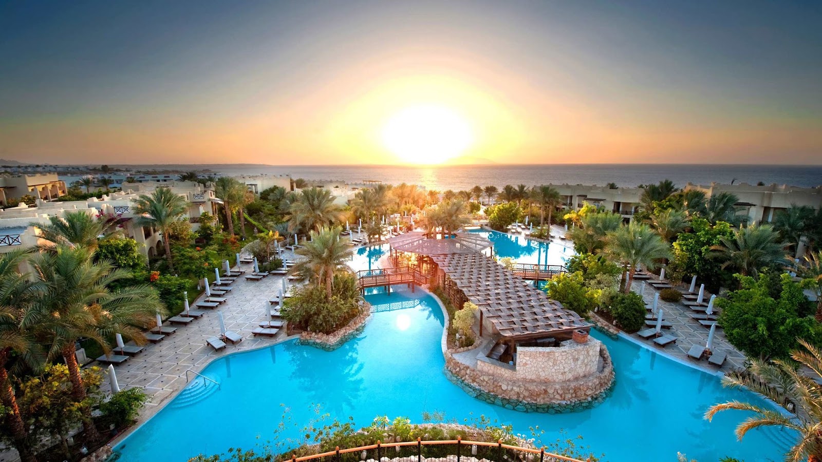 Aerial view of The Grand Hotel Sharm El Sheikh's pool complex at sunset, with palm trees, loungers, and the Red Sea visible beyond.