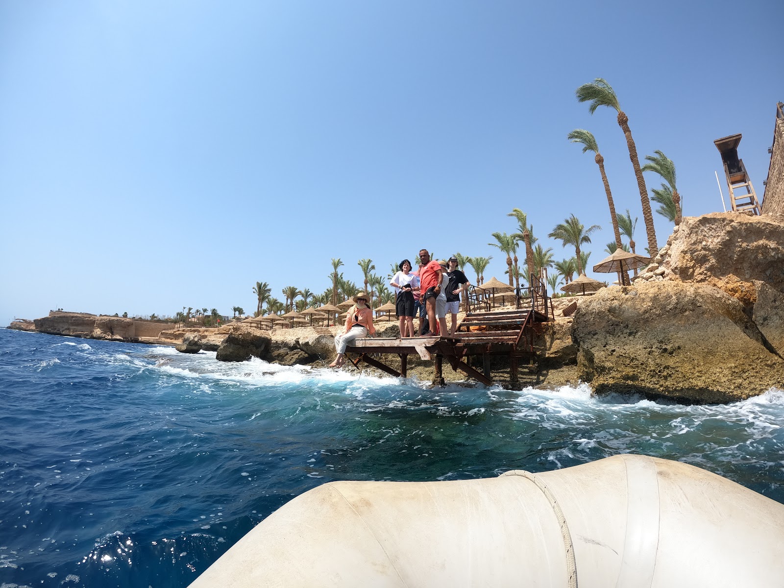 Wooden boardwalk lined with white umbrellas and loungers leads through palm-dotted grounds toward stone buildings and the Red Sea at The Grand Hotel Sharm El Sheikh.