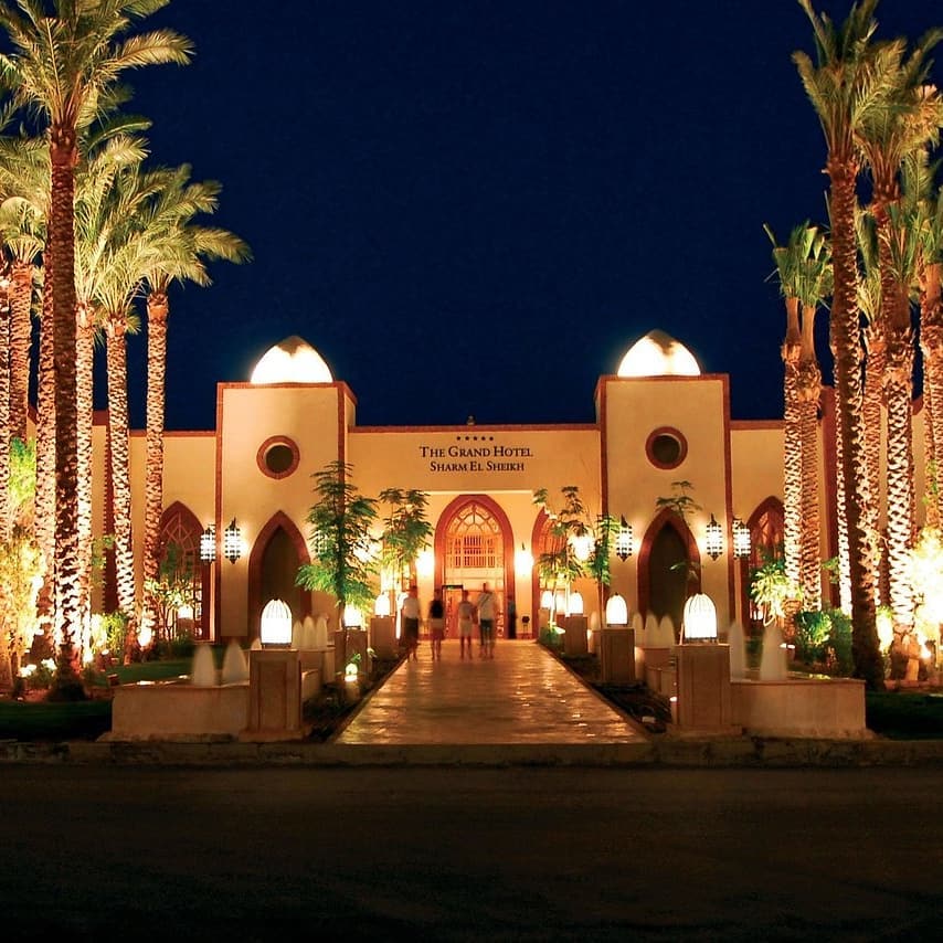 Illuminated Moorish-style building with arched windows and ornamental doors at night, flanked by tall palm trees and climbing ivy, at The Grand Hotel Sharm El Sheikh.
