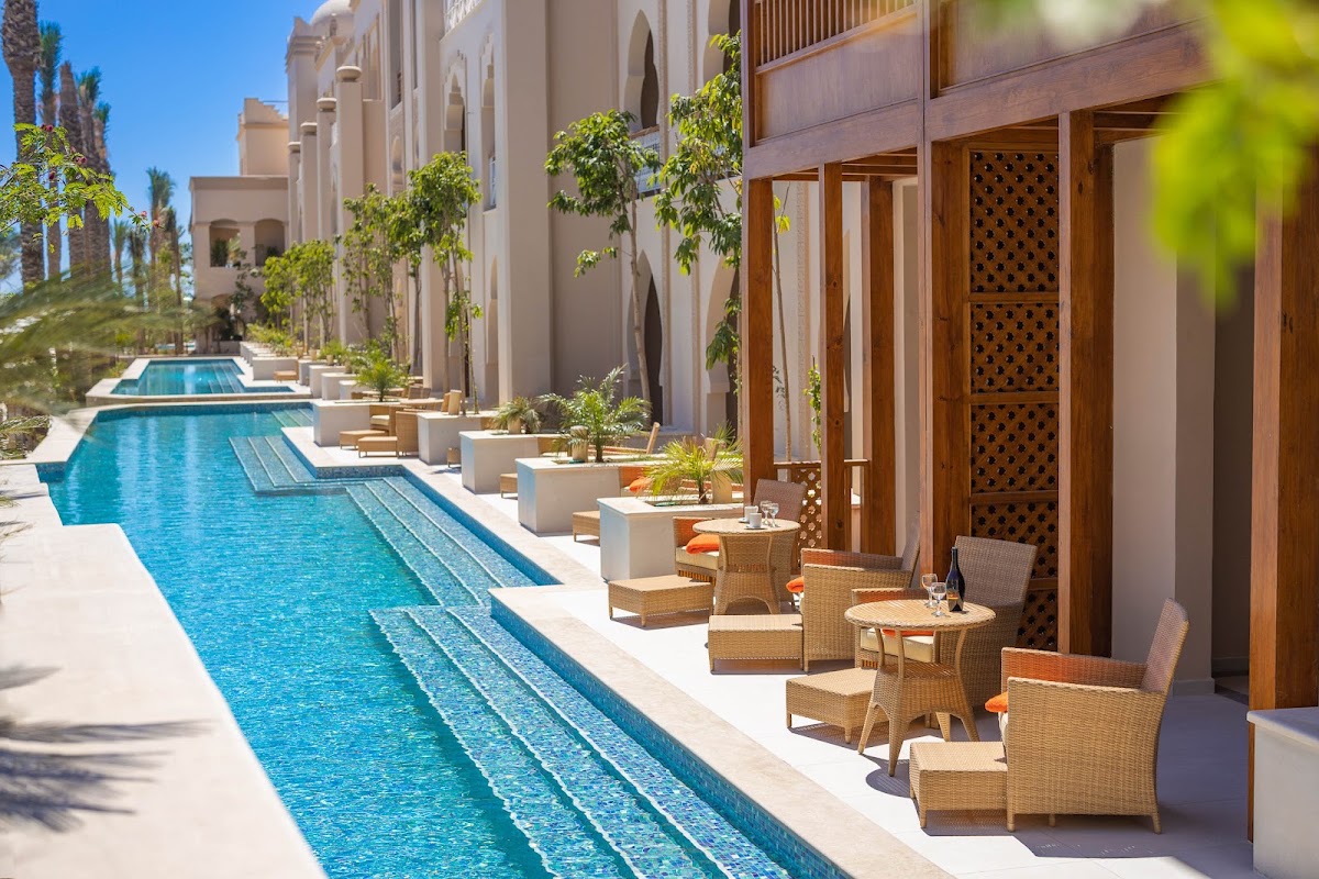 Linear pool runs alongside beige resort buildings with wooden doors, wicker seating, and climbing vines under clear blue sky.