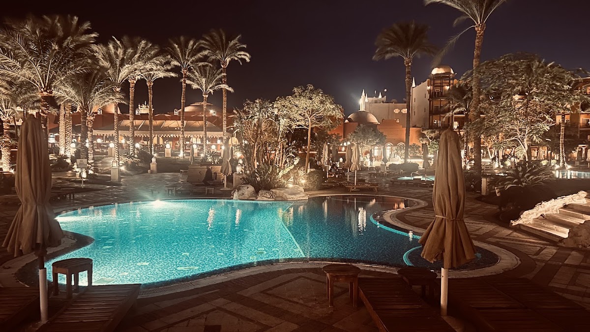 Illuminated pool at night surrounded by palm trees, lounge chairs with tan umbrellas, and resort buildings in the background at The Grand Resort.