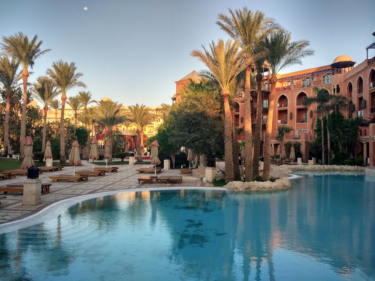 Pool area at sunset with terracotta resort buildings, palm trees, lounge chairs, and beach umbrellas at The Grand Resort Hurghada.