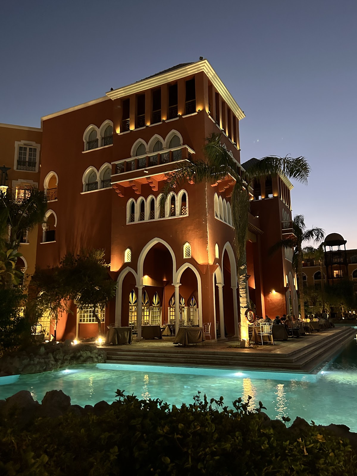 Terracotta building with arched windows and cream trim lit at dusk, reflected in an illuminated turquoise pool below, surrounded by palm trees.