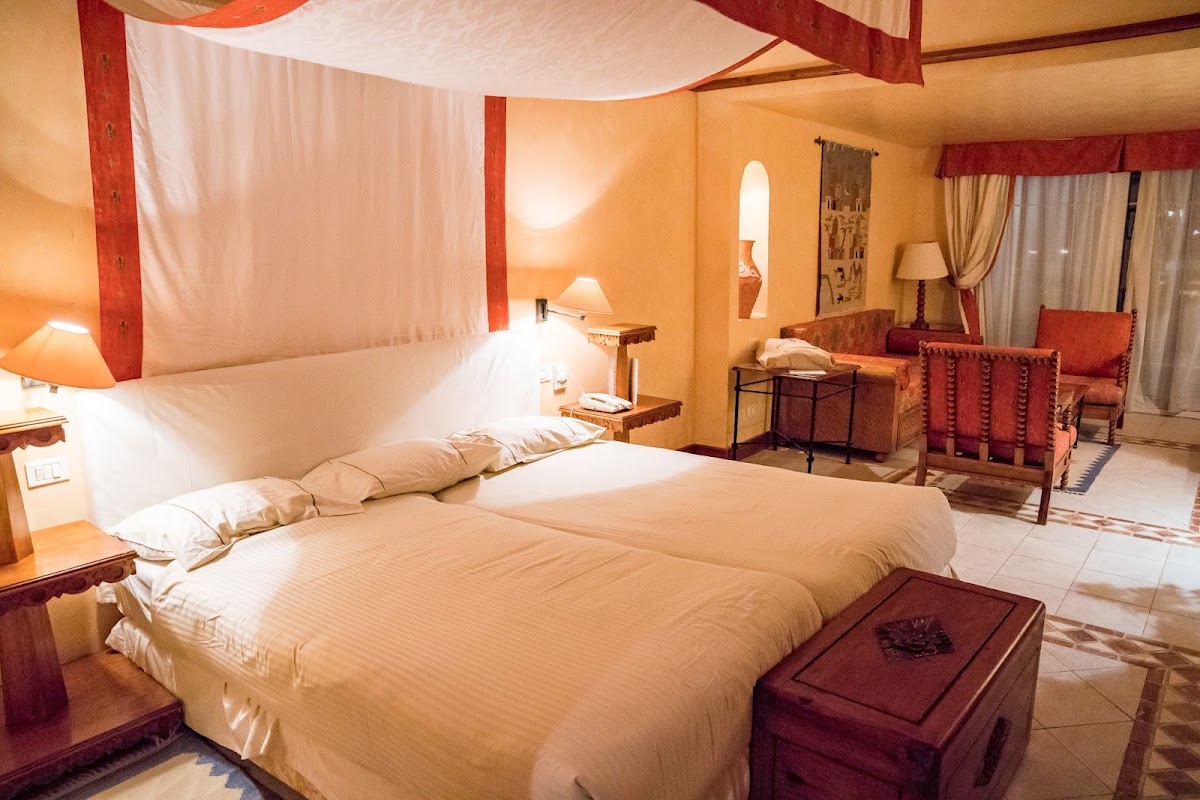 Resort suite with canopied bed, warm orange walls, red and white drapery, sitting area with upholstered chairs, and tiled flooring.
