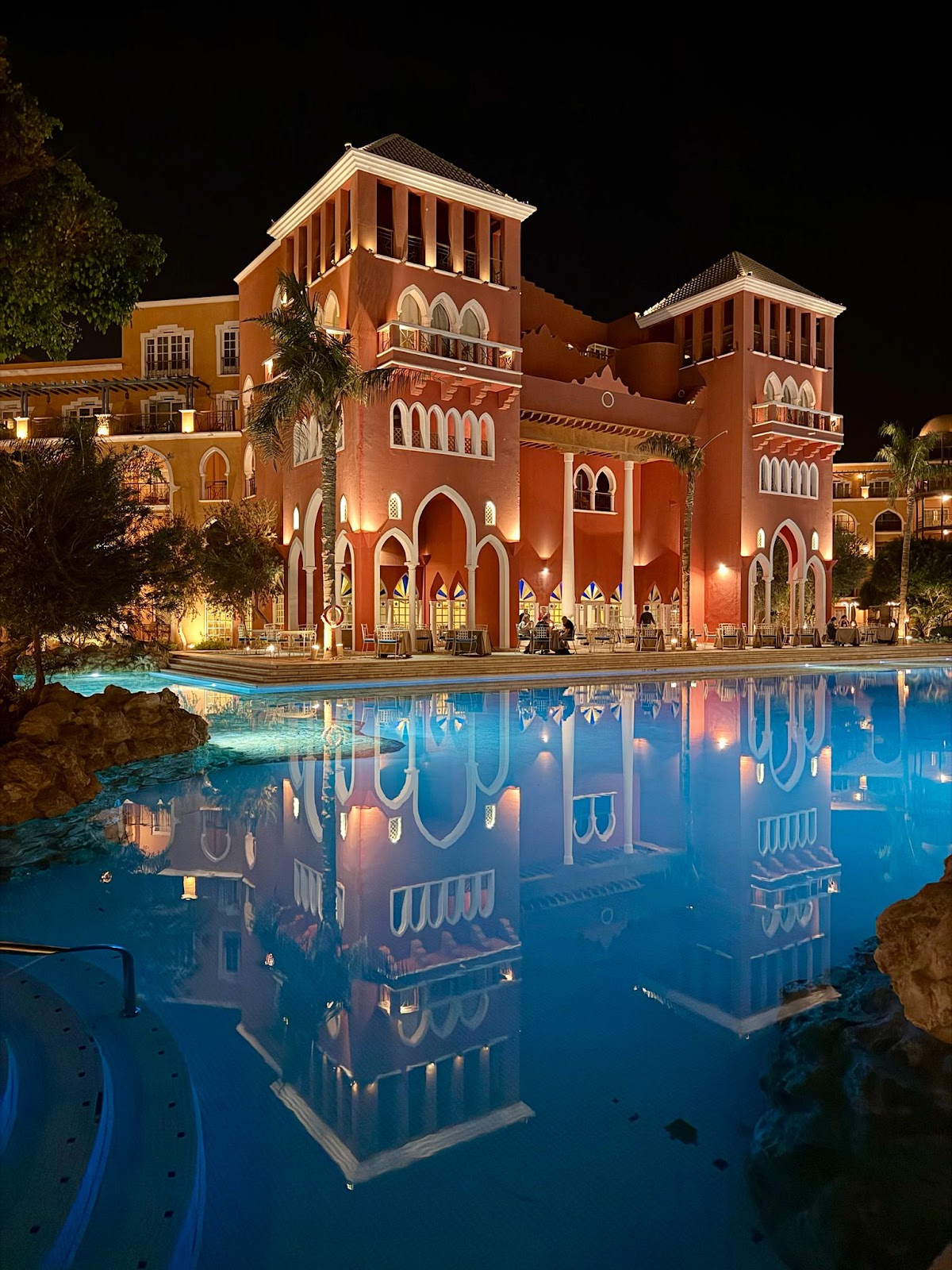 Illuminated red-brick resort buildings with Moorish architecture reflected in a glowing blue lagoon pool at night.