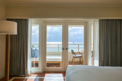 Bedroom with ocean view through open glass doors leading to a balcony with wooden chair at The House, Barbados.