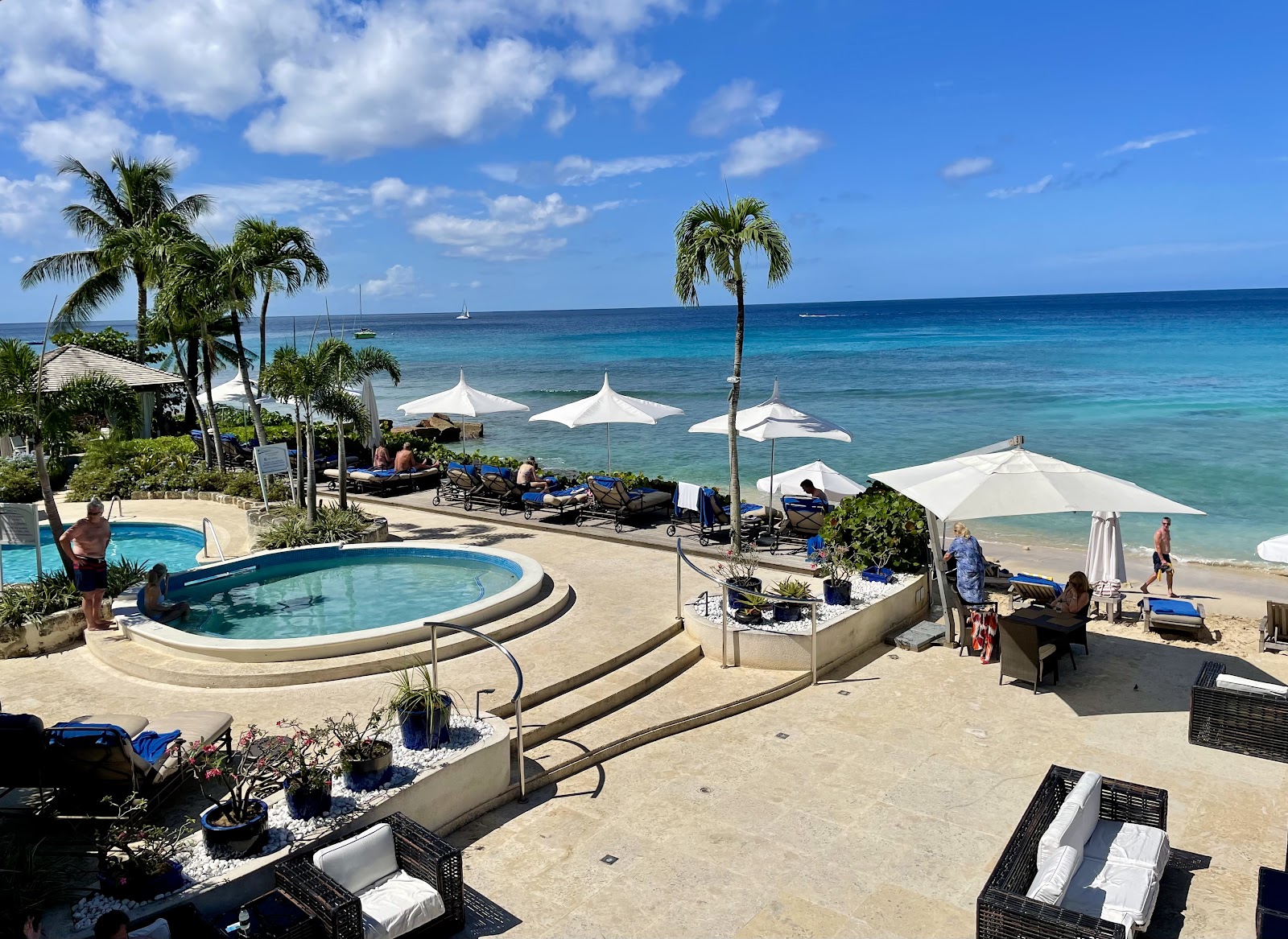 Circular pool and white umbrellas on sandy beach at The House Barbados, with turquoise ocean and sailboats beyond.