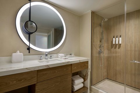 Modern bathroom with illuminated circular mirror, wooden vanity, white sink, and walk-in shower with wood-look tile walls at The House, Barbados.