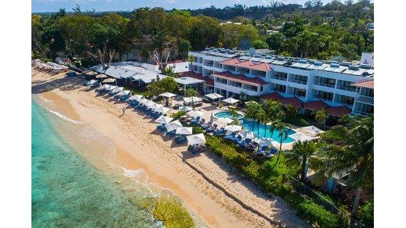 The House Barbados sits beachfront with turquoise waters, sandy shore lined with white loungers, and a blue-roofed multi-story building nestled among tropical trees.