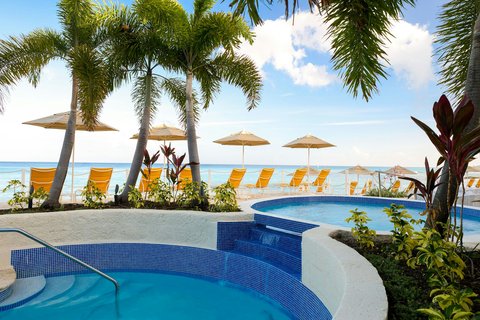 Curved blue pool with palm trees, umbrellas, and ocean view at The House resort in Barbados.