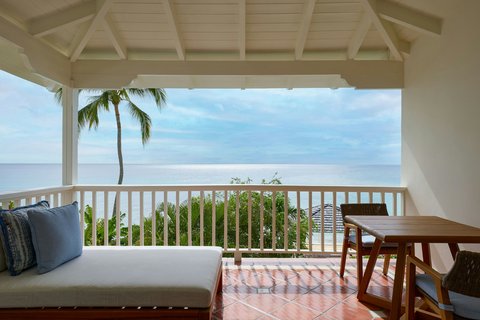 A covered oceanfront balcony at The House Barbados with a cushioned daybed, wooden dining table, and views of palm trees and Caribbean Sea.