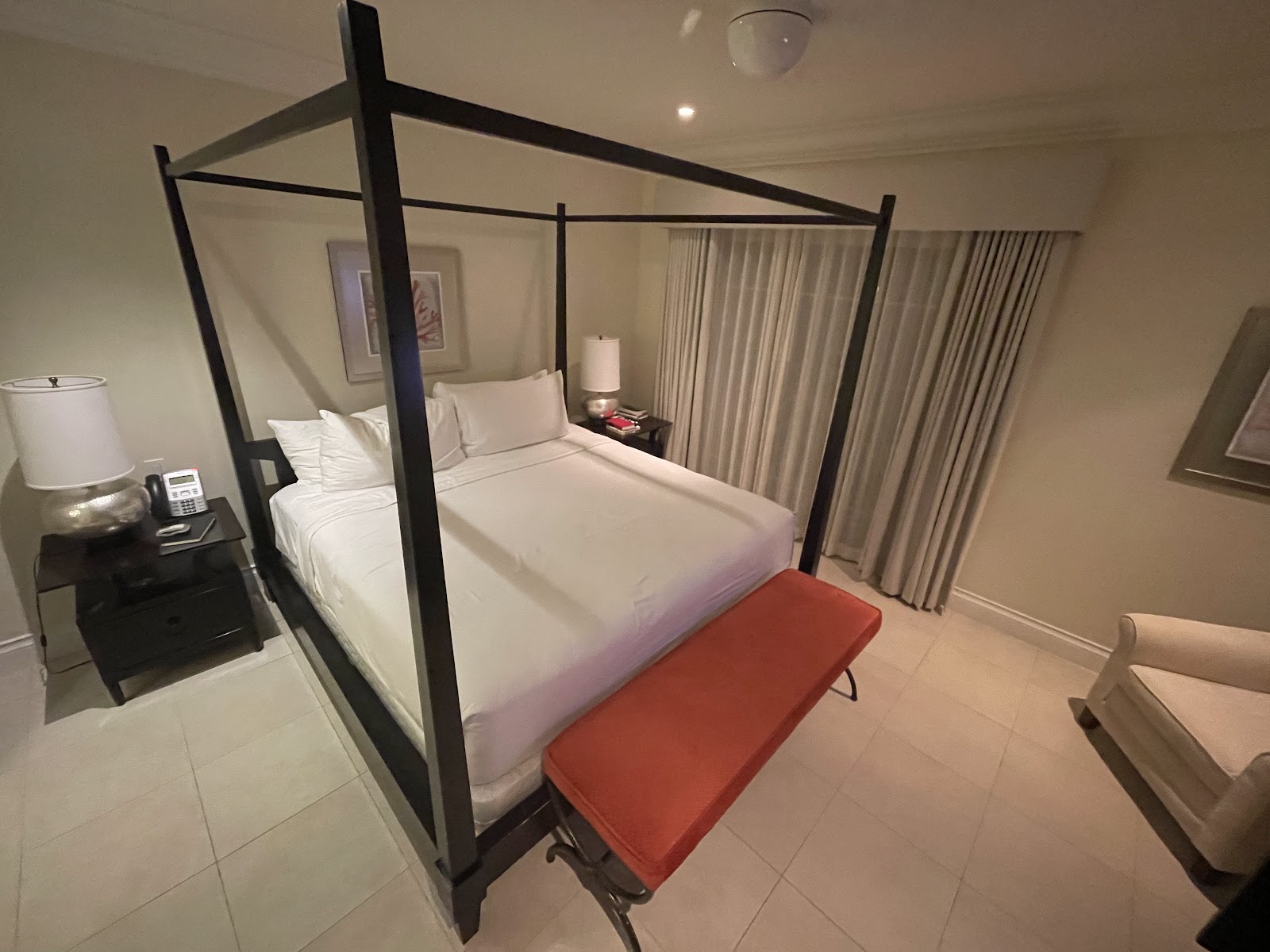Modern resort bedroom with a black four-poster bed, white linens, red bench, nightstand lamp, and cream curtains.