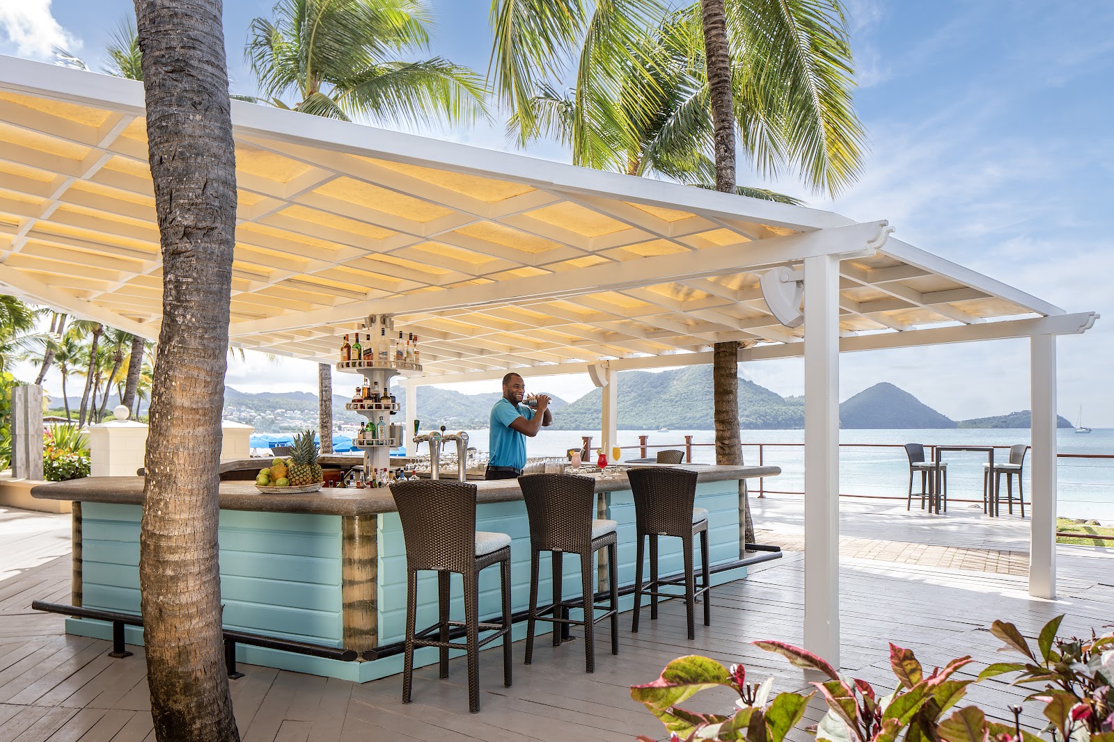 Covered waterfront terrace with wicker seating, blue umbrellas on the beach, and sailboats on the bay at sunset with mountains beyond.