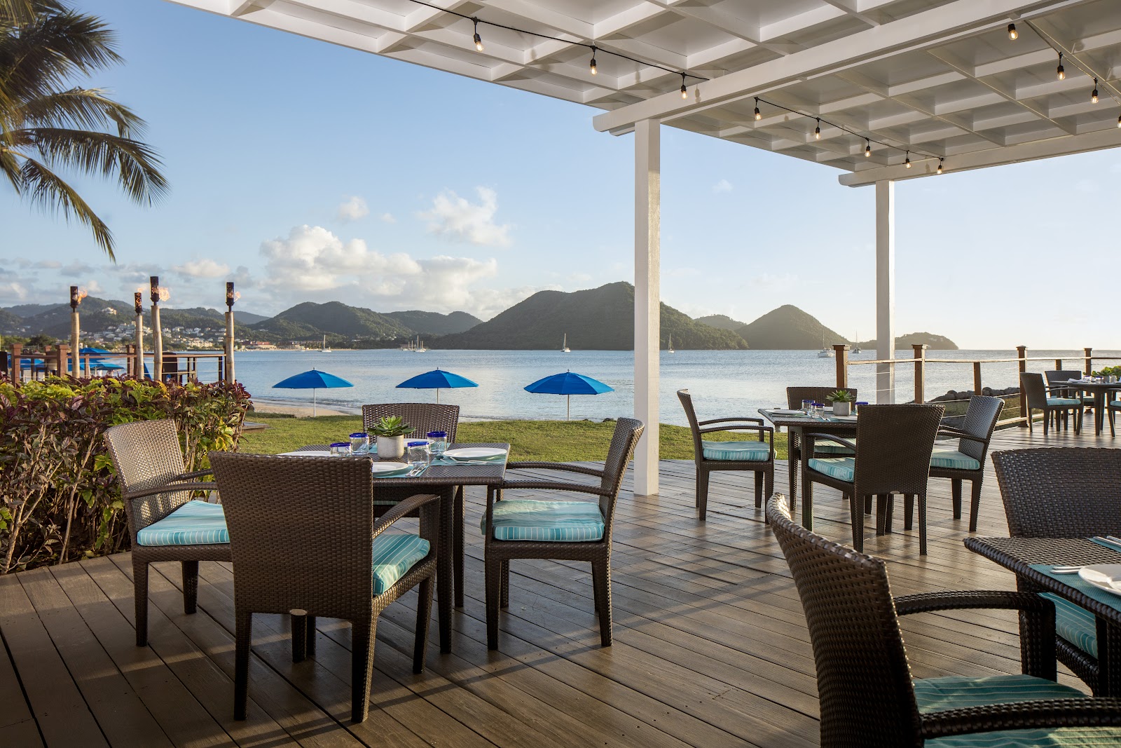 Beachfront bar with turquoise counter under white pergola, bartender pouring drinks, palm trees, and mountainous coastline views at The Landings Resort and Spa.