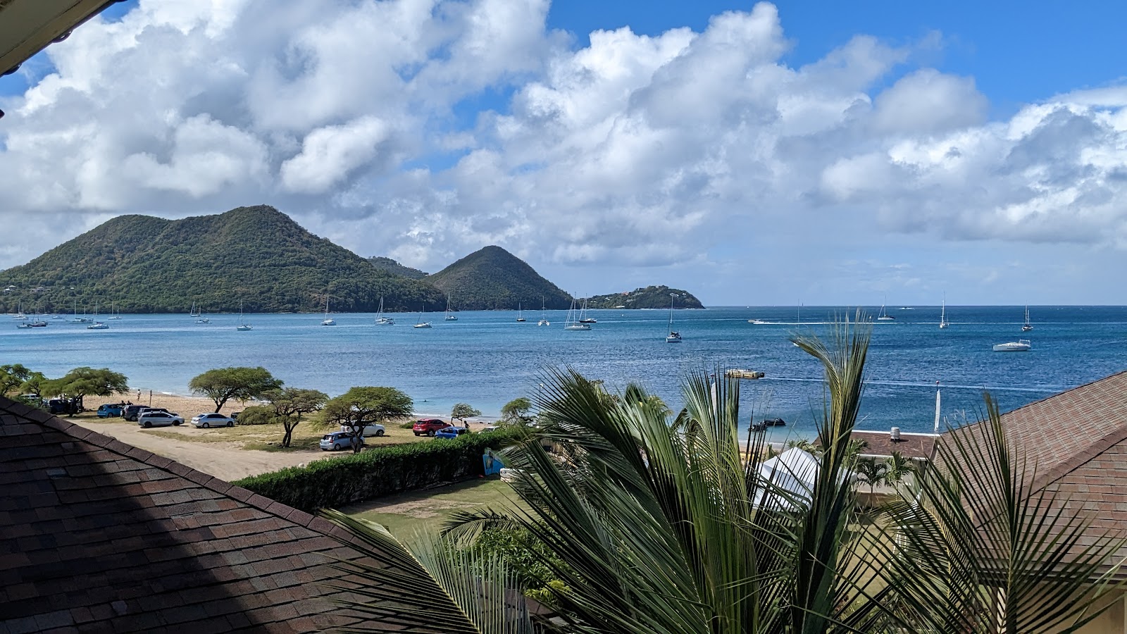 Palm fronds frame a view of Pigeon Island Bay with sailboats anchored in turquoise water and forested peaks rising beyond the beach at The Landings Resort and Spa.